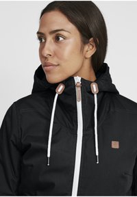 Black hooded jacket with white zipper, featuring tan accents and drawstrings. The material has a smooth texture and simple, functional design.