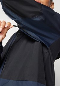 Marineblau und schwarz zweifarbiger Windbreaker mit hohem Kragen, Reißverschluss und glatter Textur. Verfügt über eine Kapuze und verstellbare Kordelzüge.