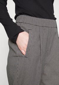 Hand in pocket of houndstooth-patterned pants paired with a black long-sleeve top, showing fabric texture and stitching details.