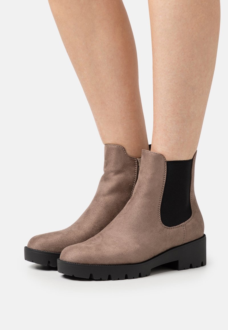 Bottines en daim marron avec panneaux latéraux élastiques noirs et semelles épaisses noires portées sur jambes nues sur fond blanc.