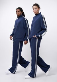Navy blue tracksuits with white vertical stripes, featuring zip-up jackets and wide-leg pants. Made of soft fabric, minimalistic design.