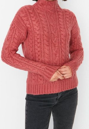 Femme portant un pull à col roulé rose en maille torsadée à manches longues et un jean noir, les mains jointes devant.
