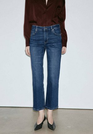 Donna che indossa jeans blu cropped a gamba dritta, camicia a maniche lunghe rosso scuro e tacchi neri a punta, in piedi davanti a uno sfondo uniforme.