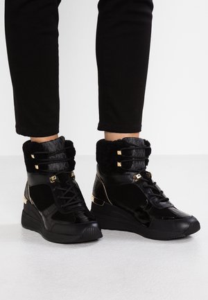 Baskets compensées noires avec col en fourrure et accents dorés portées avec un pantalon noir sur un sol blanc.