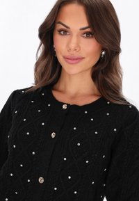 Schwarze Strickjacke mit strukturiertem Muster, verziert mit silbernen Perlen und versehen mit runden, metallischen Knöpfen.