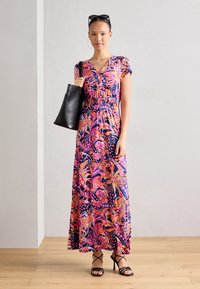 Robe maxi colorée et à motifs avec des manches courtes, décolleté en v et taille cintrée. Le mannequin tient un sac fourre-tout noir et porte des talons aiguilles noirs.