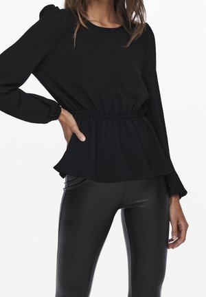 Schwarzes Langarmoberteil mit gerafftem Taillenbund, strukturiertem Stoff und dezenten Puffschultern, kombiniert mit glänzenden schwarzen Leggings.