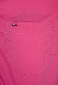 Rosa Baumwollstoff mit einer einzigen Gesäßtasche. Die Tasche verfügt über ein kleines gesticktes Logo in Rot, Weiß und Blau. Glatte Textur.