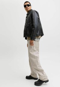 Άνδρας που φοράει μαύρο δερμάτινο μπουφάν, μπεζ παντελόνι, μαύρα παπούτσια, γυαλιά ηλίου, στέκεται μπροστά σε απλό ανοιχτό φόντο.