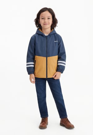 Kind met schouderlang donker haar, draagt een marineblauwe en mosterdkleurige jas met capuchon, blauwe spijkerbroek en bruine laarzen, staand en glimlachend.