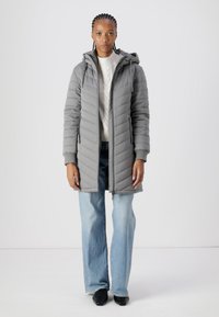 Polstret grå frakk med hette, med glidelås foran, diagonalt kviltet mønster og sidelommer; lagd over en kremfarget genser og blå jeans.