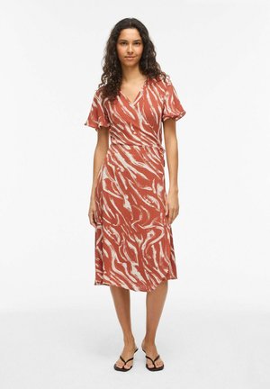 Femme aux longs cheveux bouclés portant une robe portefeuille midi à motifs rouille et blancs et des sandales noires, debout devant un fond blanc uni.
