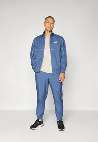 Mėlynas sportinis kostiumas iš lengvos medžiagos su baltais akcentais, sudarytas iš striukės su užtrauktuku ir elastinių rankogalių, derinamas su juodais sportiniais bateliais.