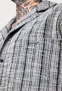 Grau und schwarz gestreiftes Button-Up-Hemd aus strukturiertem Stoff, mit einer Brusttasche und einem "ESTEEM"-Metalltag. 