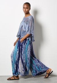 Top all'uncinetto blu chiaro con maniche larghe, abbinato a pantaloni a gamba larga con motivo blu e verde. Sandali neri flat completano l'outfit.