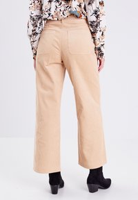 Pantalon beige en velours côtelé à jambes larges avec tissu texturé, deux poches arrière et une taille haute, associé à des bottes noires à talons.