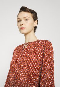 Κόκκινο φόρεμα με σχέδιο, με σχήμα κλειδώματος στον λαιμό, μακριά μανίκια και ελαστική μέση. Διαθέτει λουλουδάτα και γεωμετρικά σχέδια σε αντίθετες αποχρώσεις.