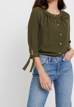 Kobieta w oliwkowozielonej bluzce na guziki z wiązanymi rozcięciami na rękawach i jasnoniebieskich, wysokowysokościowych dżinsach, stojąca z jedną ręką w kieszeni.
