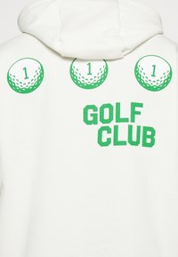 Vit hoodie med grön grafisk tryck. Tre golfbollsmotiv med siffran '1' ovanför, och "GOLF KLUBB" tryckt med fet grön text.