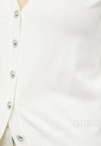 Vit cardigan i mjukt, strukturerat tyg med silverfärgad knappar och en broderad logotyp. Klassisk design med ribbade detaljer.