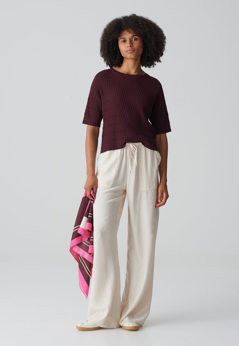 Burgunderfarbenes, geripptes Kurzarmoberteil mit abgerundetem Saum, kombiniert mit cremefarbenen, weiten Hosen. Hält einen rosa und braun gemusterten Schal.