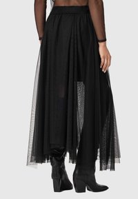 Jupe maxi en tulle noir avec un design superposé, dotée d'une taille ajustée et d'une texture transparente, associée à des bottes noires montée jusqu'aux genoux.