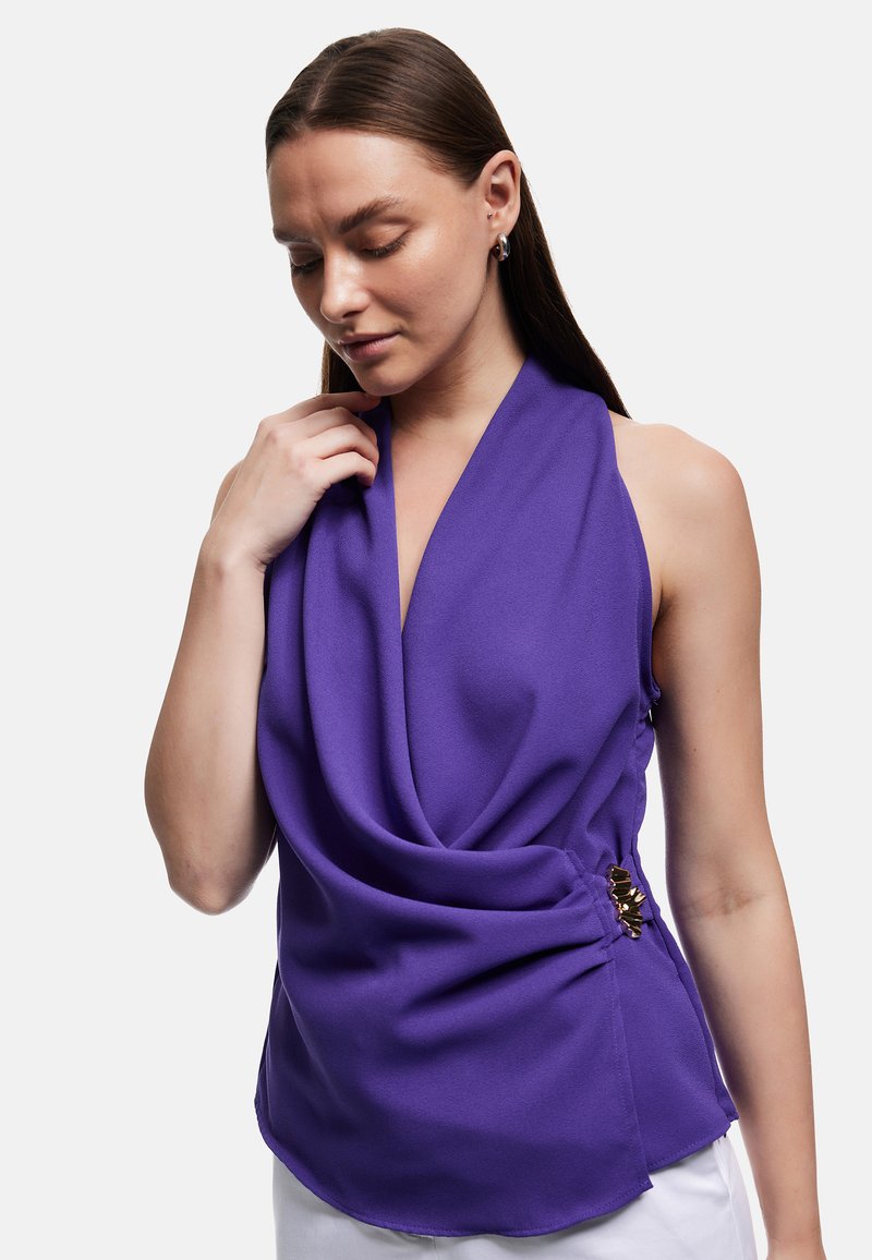 Femme aux cheveux bruns et lisses portant un haut violet sans manches drapé avec une boucle dorée sur le côté, regardant vers le bas et touchant l'encolure.