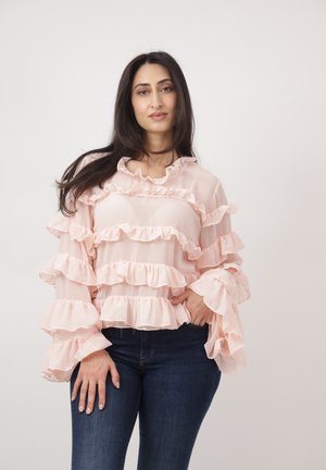 Vrouw met lang donker haar, gekleed in een lichtroze blouse met ruches en donkerblauwe spijkerbroek, staand voor een eenvoudige lichte achtergrond.