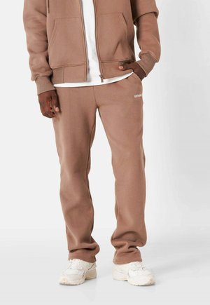 Brune sweatpants med en afslappet pasform, der har et lille hvidt logo på venstre side. Sættes sammen med en matchende zip-up hættetrøje og hvide sneakers.