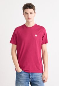 Burgundy színű pamut póló rövid ujjakkal és kerek nyakkal; az elején egy kis fehér logó folt található.