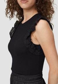 Zwarte mouwloze top met geribbeld textuur, voorzien van decoratieve zwart kant ruffles op de schouders, gecombineerd met een donkere geplooide rok.