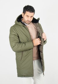 Manteau matelassé vert olive avec capuche, doté d'une fermeture éclair sur le devant et de poches latérales. Intérieur doublé en marron foncé, texture lisse avec des détails de couture.