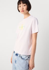 Lys rosa bomull t-skjorte med en gul og grønn Calvin Klein-logo på brystet, rund halslinning og korte ermer.