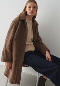 Manteau teddy marron avec un col et des boutons, porté sur un pull beige clair et un pantalon large noir. Texture lisse.