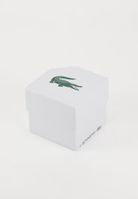Boîte en carton blanc avec une surface texturée, arborant un logo de crocodile vert et le mot "LACOSTE" sur le côté. Design à dessus fermé.