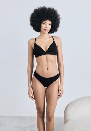 Γυναίκα με αφρο χτένισμα, που φοράει μαύρο σουτιέν και εσώρουχο, στέκεται δίπλα σε μια λευκή υφή πολυθρόνα πάνω σε ανοιχτό γκρι φόντο.