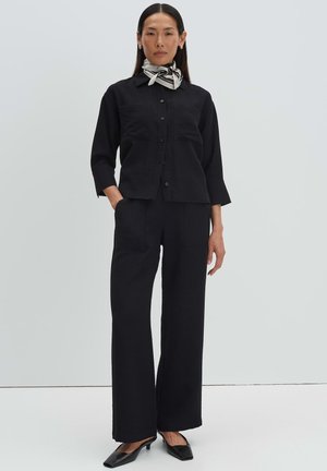Femme debout portant une chemise noire boutonnée, un pantalon noir à jambes larges, des talons noirs et un foulard blanc à motif autour du cou sur un fond uni.