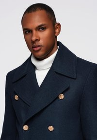 Marineblauer Woll-Peacoat mit einem doppelten Knopfdesign, großen Revers und goldenen Knopfakzenten, kombiniert mit einem weißen gerippten Rollkragenpullover.
