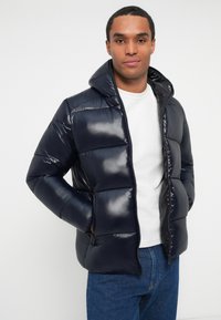 Navy puffer jacket with a shiny finish, zip closure, hood, and horizontal quilted design. Worn over a white shirt and paired with blue jeans.