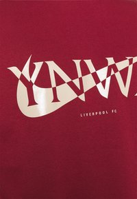 Tissu rouge avec de grandes lettres stylisées de couleur crème sur lesquelles se superpose un design en forme de virgule, avec un petit texte indiquant « Liverpool FC » en dessous.