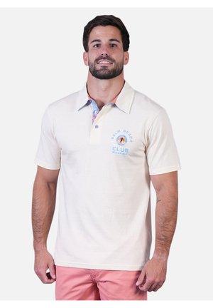 Homme portant un polo blanc avec le logo "Palm Beach Club" et un short rose, debout devant un fond blanc uni.
