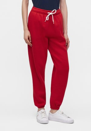 Model iført knaldrøde joggers med snøre og elastiske manchetter samt hvide sneakers, stående foran en ensfarvet baggrund.