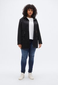 Μαύρο αδιάβροχο μπουφάν με κουκούλα, κλείσιμο με φερμούαρ και πλαϊνές τσέπες. Συνδυασμένο με λευκό μπλουζάκι και μπλε τζιν, φορεμένο με μπεζ αθλητικά παπούτσια.