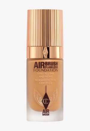 Foundation in einer klaren Glasflasche mit einem goldenen geriffelten Deckel. Das Etikett zeigt braune Schrift und ein geflügeltes Design. Die Farbe ist ein warmer Bronzeton.