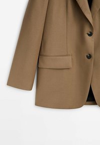 Brun enkeltstående blazer med to sorte knapper, lange ærmer og en klaplomme på venstre side.