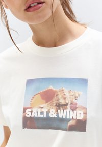 Cremefarvet bomulds T-shirt med et fremtrædende grafisk motiv af en musling med teksten "SALT & WIND" i fede hvide bogstaver nedenfor.