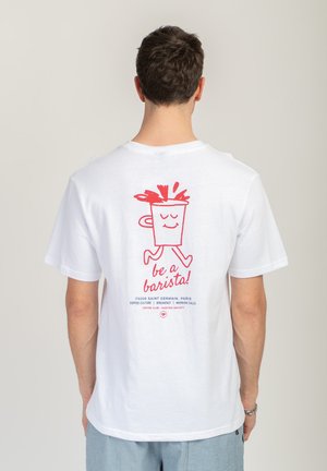 Mężczyzna w białej koszulce z czerwonym rysunkiem uśmiechniętej filiżanki kawy idzie, a na plecach ma napis „bądź baristą!”.