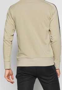 Uomo che indossa una felpa beige con righe nere sulle maniche e pantaloni neri, mostrato di spalle su uno sfondo neutro.