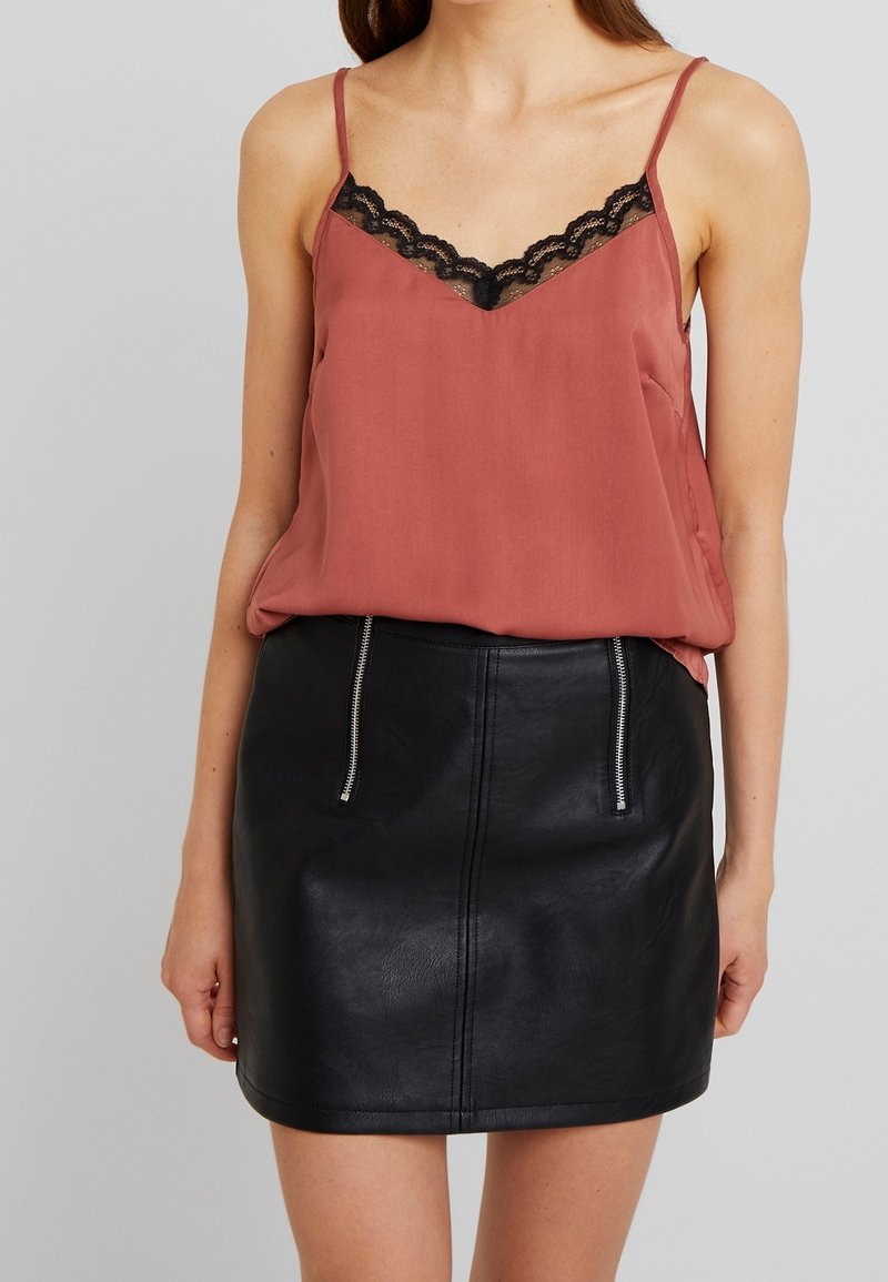 Femme portant un caraco couleur rouille avec bordure en dentelle noire et une mini-jupe en cuir noire avec poches zippées à l'avant.