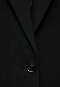 Černý blazer s klopou, texturovanou látkou a uzávěrem na jeden černý knoflík. Detailní pohled zdůrazňuje detaily a švy.